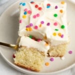A closeup of a slice of vanilla cake topped with creamy white frosting and colorful confetti sprinkles, with a bite already on a gold fork.
