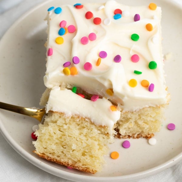 A closeup of a slice of vanilla cake topped with creamy white frosting and colorful confetti sprinkles, with a bite already on a gold fork.