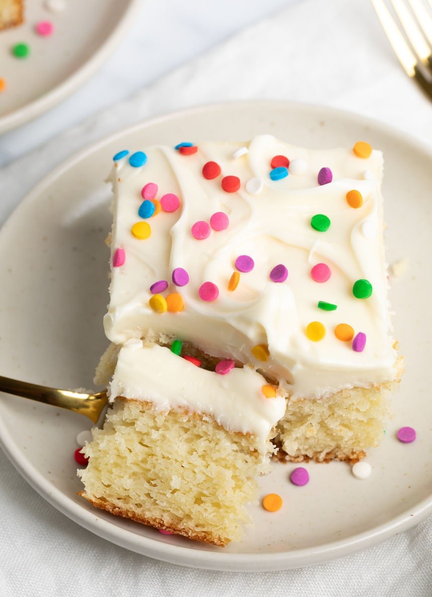 A tender slice of yellow cake with fluffy white dairy free frosting and bright round sprinkles on a white plate with a bite on a gold fork that is almost out of frame