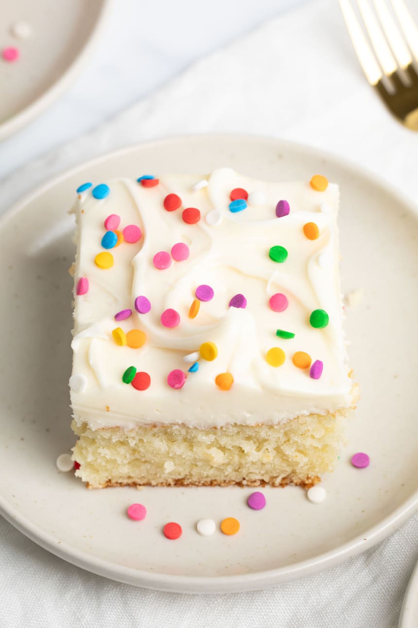 A square slice of vanilla-frosted cake with sprinkles, served on a cream-colored plate on a white cloth