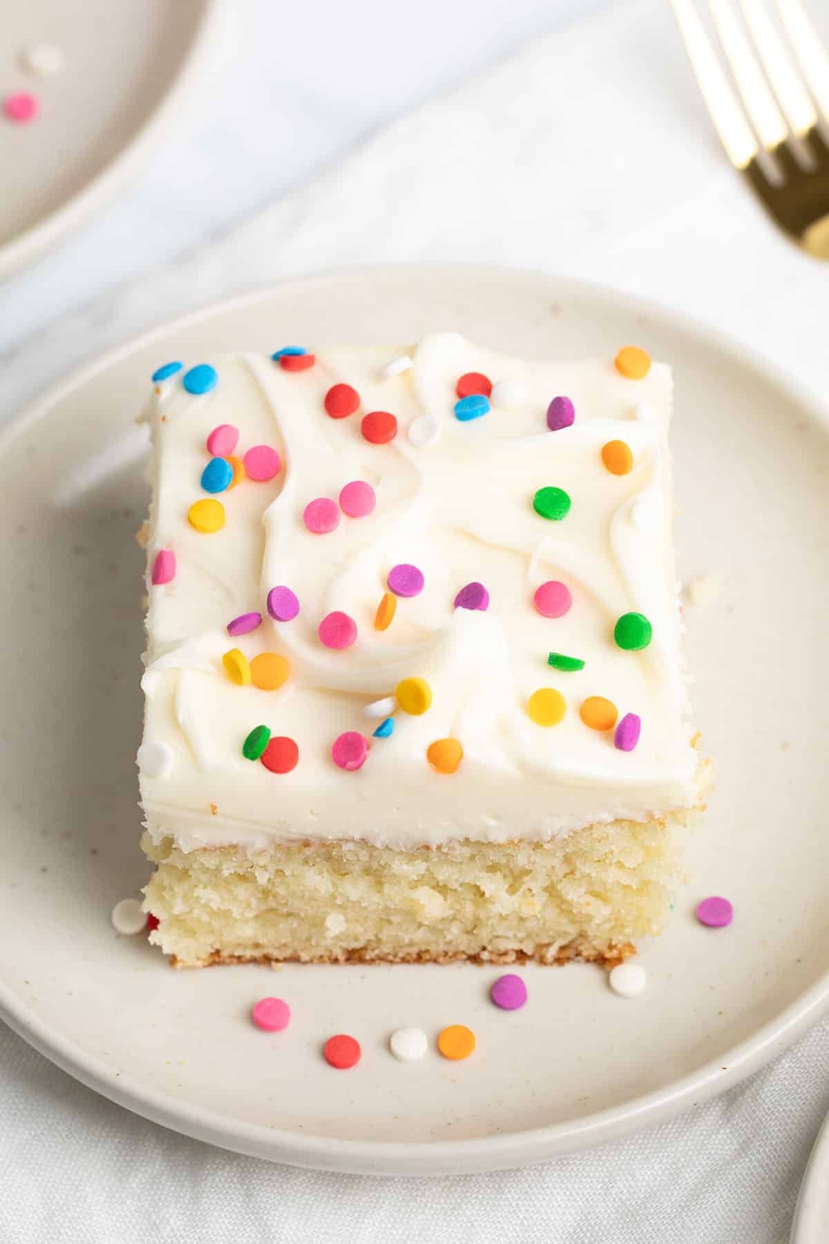 A square slice of vanilla-frosted cake with sprinkles, served on a cream-colored plate on a white cloth