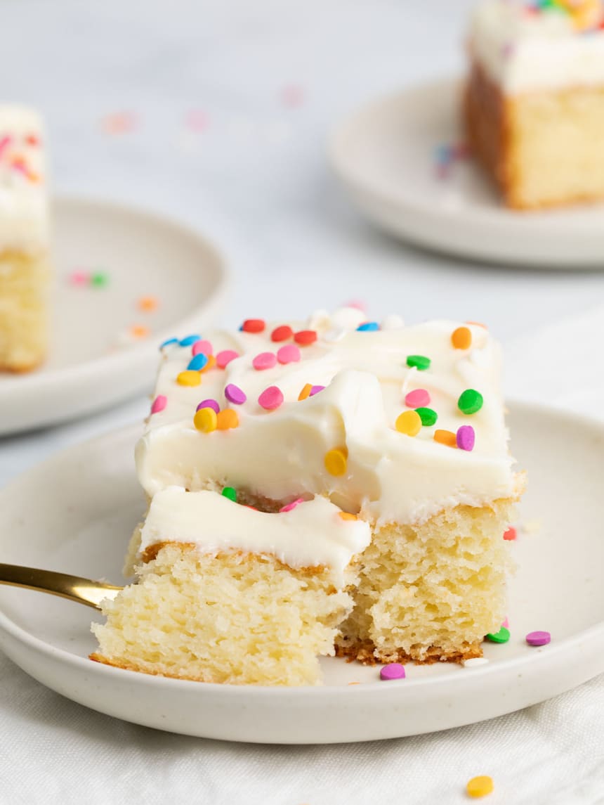 A forkful taken out of a slice of moist vanilla cake with a thick layer of frosting and sprinkles on a small round plate with two other plates and slices in the background