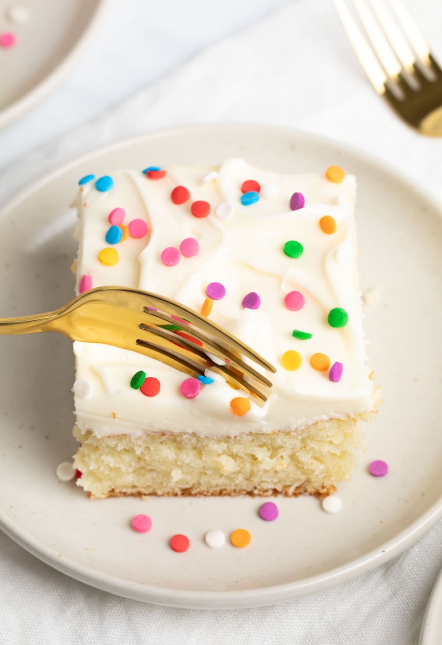 Golden fork poised above a slice of frosted vanilla cake, pressing gently into the soft frosting above the tender crumb of the cake