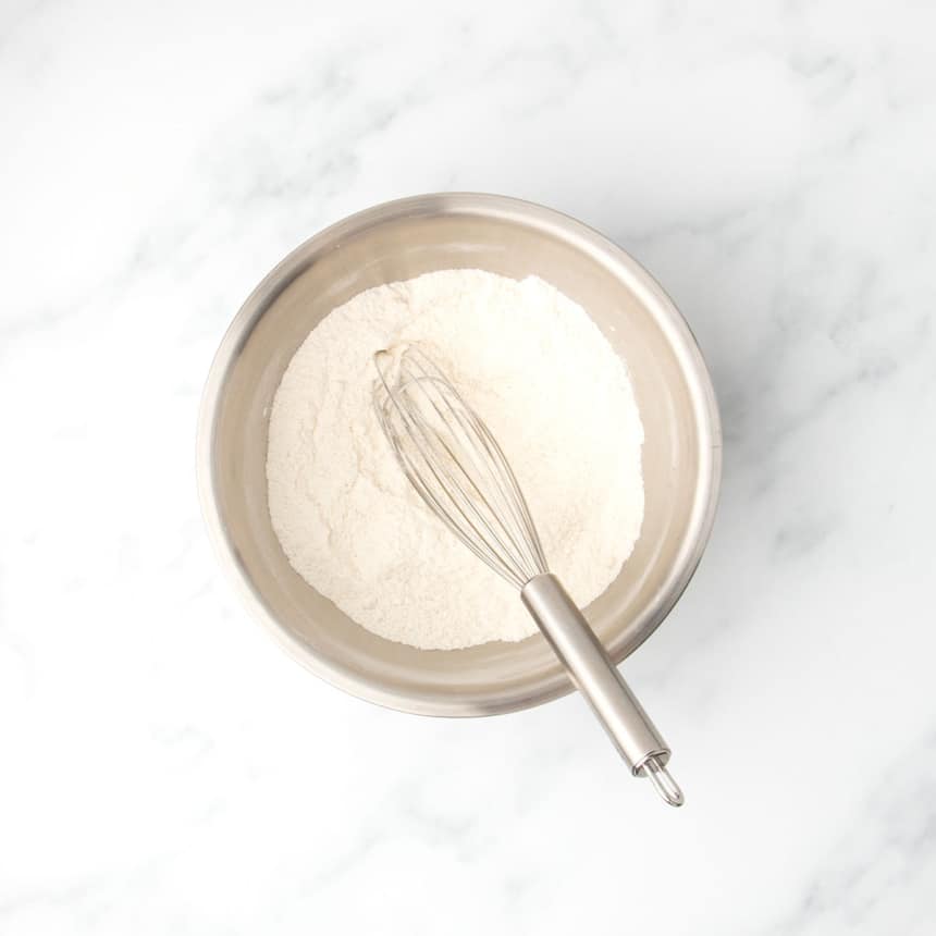 Metal mixing bowl filled with gluten free flour blend and other white dry ingredients and a stainless steel whisk resting inside, set on a white marble countertop.