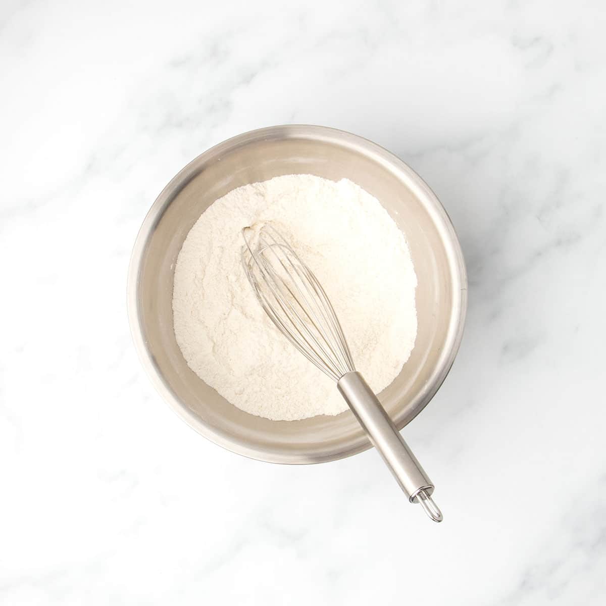 Metal mixing bowl filled with gluten free flour blend and other white dry ingredients and a stainless steel whisk resting inside, set on a white marble countertop.