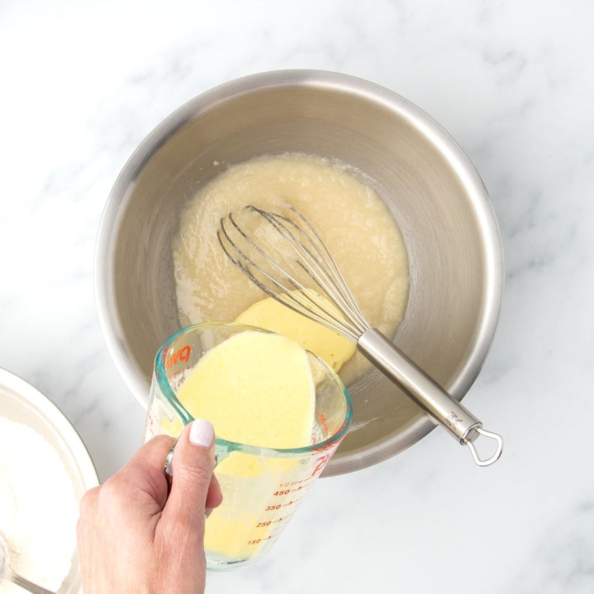 Wet ingredients being poured into the oil and sugar mixture with some dry ingredients incorporated in a large bowl, with a metal whisk resting inside.