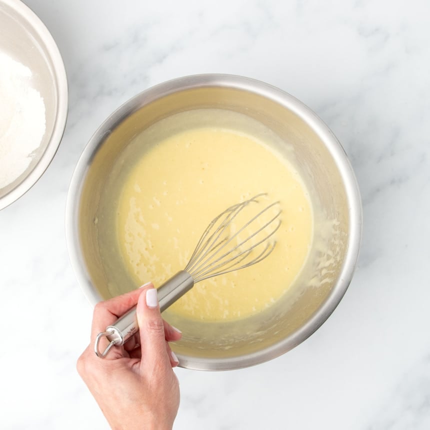 Same metal mixing bowl with some dry ingredients and all wet ingredients forming a pale yellow batter, with a whisk held in hand.