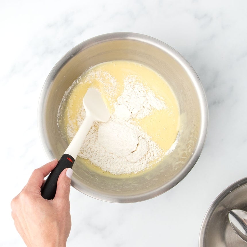 Hand holding a spatula above a bowl of cake batter, adding final dry ingredient gluten free flour mixture gradually to the rest of the batter.