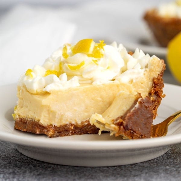 A close-up of a creamy slice of gluten free lemon pie with a golden crust, piped whipped cream, and lemon zest curls.