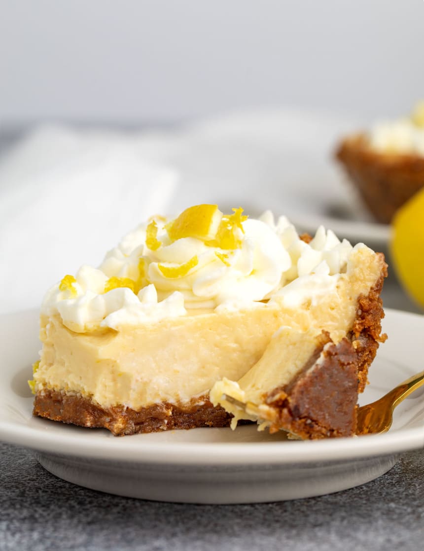 Slice of lemon pie with a cookie crust and whipped cream on a white plate, a forkful taken out to show the smooth texture