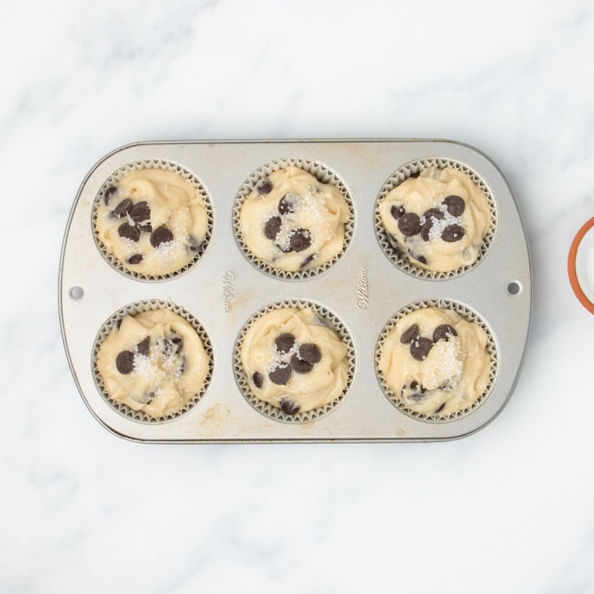 Muffin batter portioned into a 6-cup muffin tin lined with muffin liners, topped with extra chocolate chips and coarse sugar.