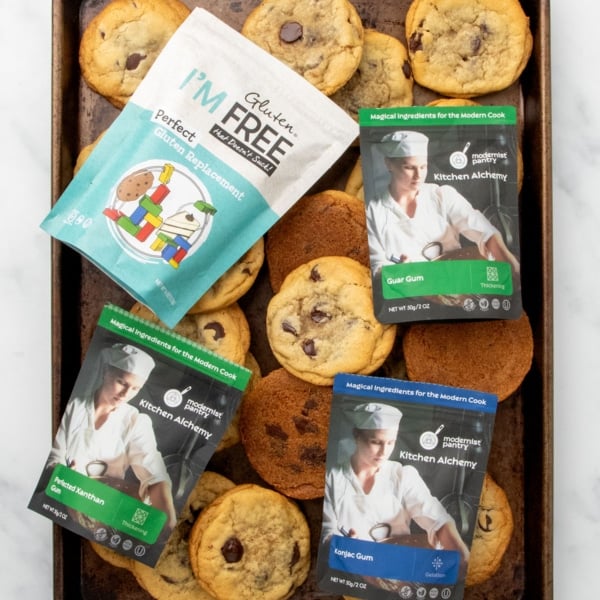 dark brown baking sheet with gluten free chocolate chip cookies and packages of xanthan gum, and xanthan gum substitutes including guar gum, konjac powder, and perfect gluten replacement on top