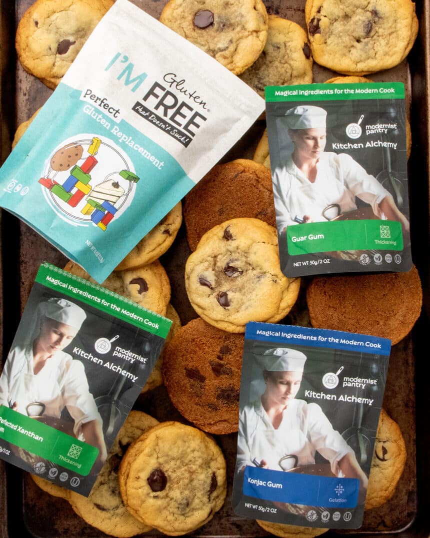 dark brown baking sheet with gluten free chocolate chip cookies and packages of xanthan gum, guar gum, konjac powder, and perfect gluten replacement on top