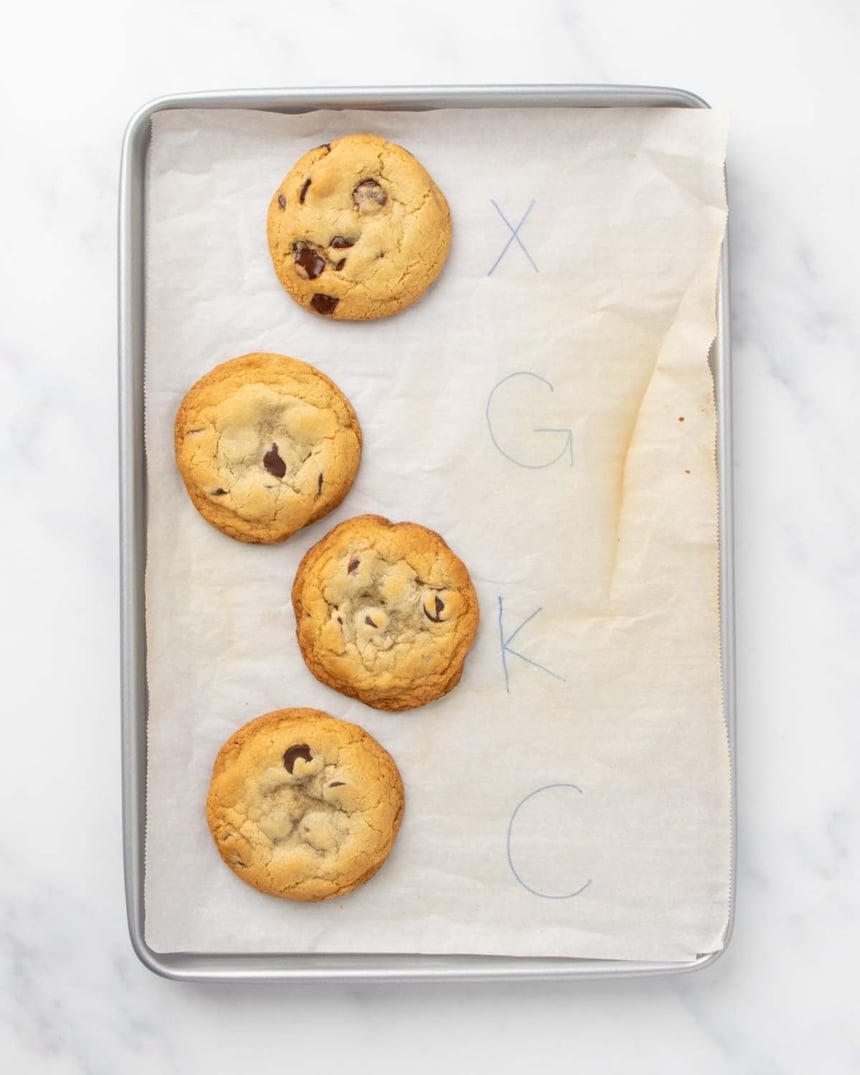 4 baked chocolate chip cookies on white parchment on baking sheet next to letters