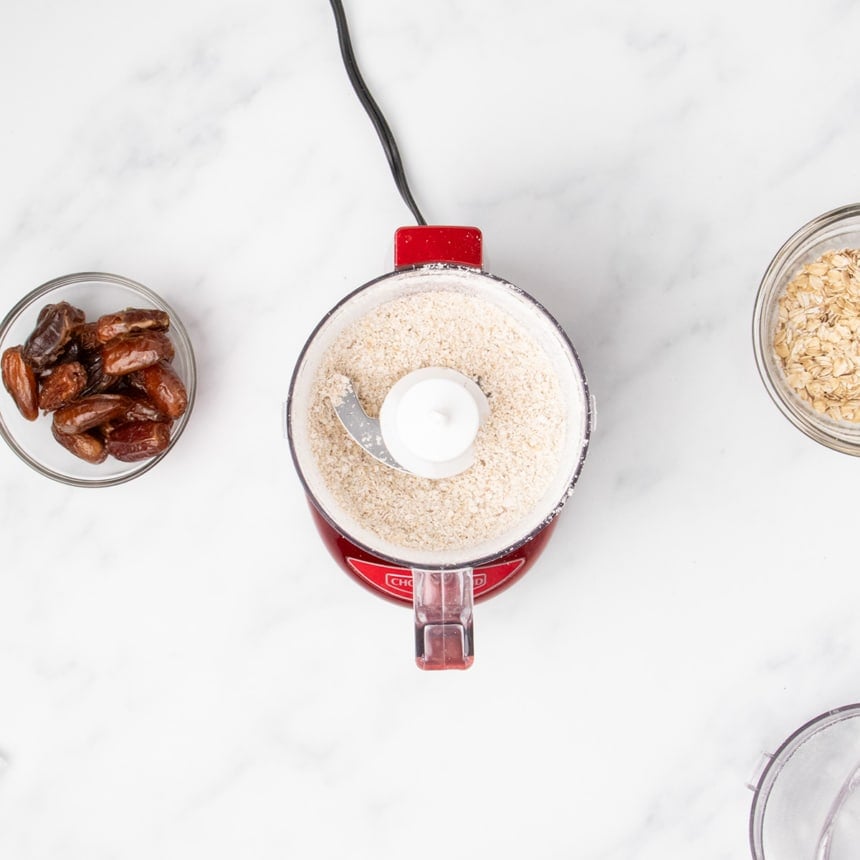 small red mini chop food processor with clear plastic bowl with ground oats, dates in a bowl on the side