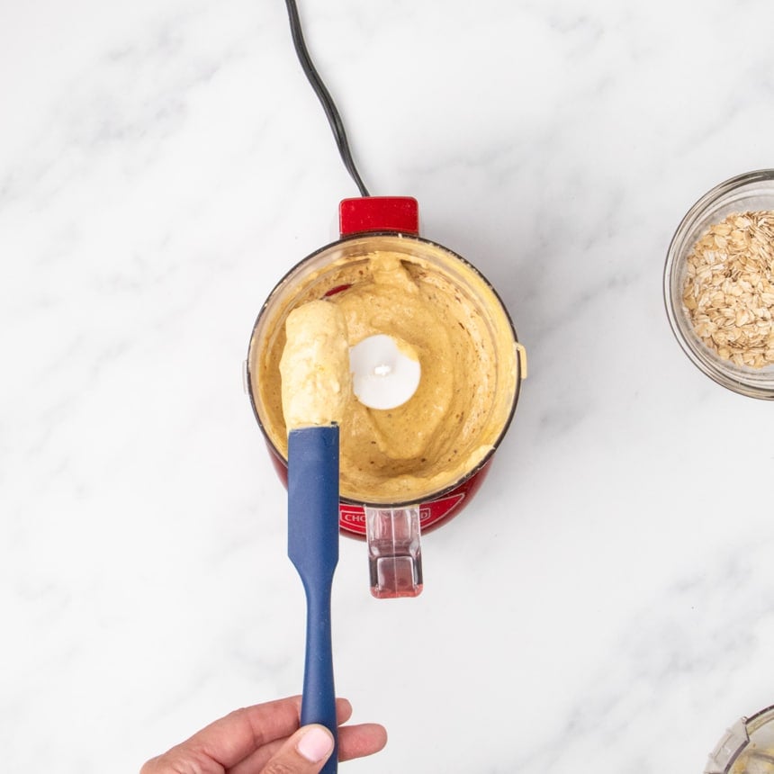 hand holding small spatula with soft light brown mixture over mini chop food processor with rest of mixture