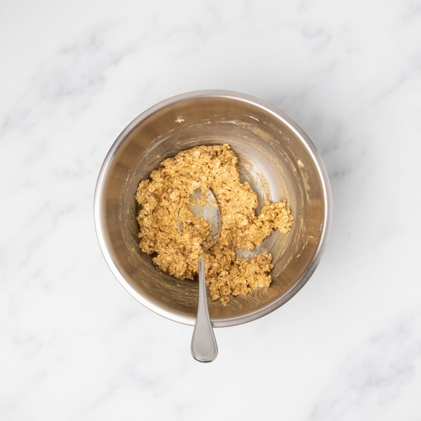 raw light brown thick raw cookie dough mixture with some rolled oats in round metal mixing bowl with large metal mixing spoon