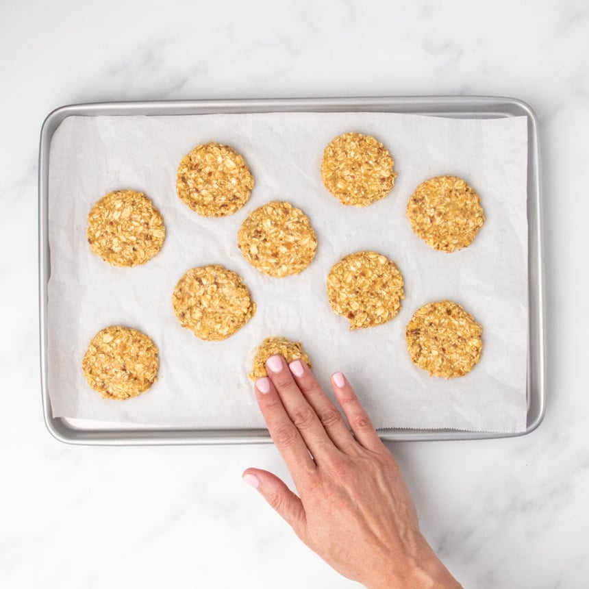 fingers pressing down on mound of light brown grainy raw cookie dough in one portion on paper lined baking sheet with 9 other shaped round raw cookies