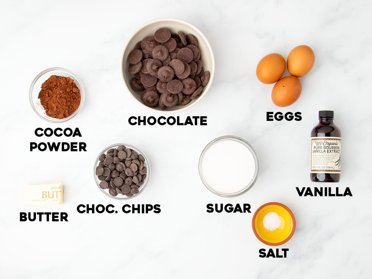 Overhead view of labeled ingredients for flourless brownies, including chocolate, cocoa, eggs, sugar, and butter