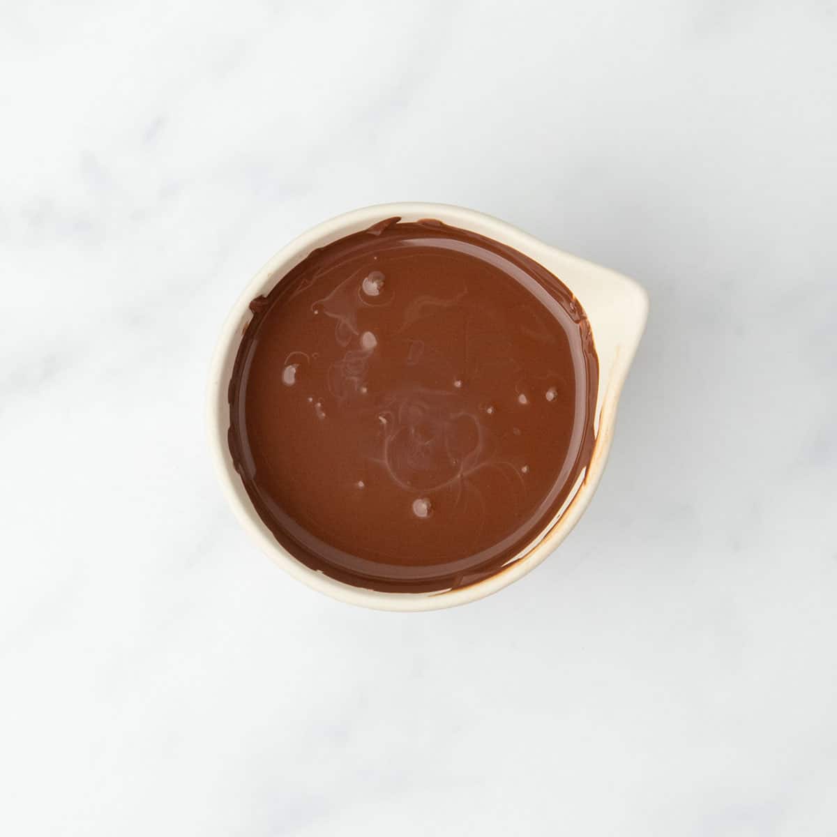 Melted dark chocolate swirled smooth in a white spouted bowl on a marble surface