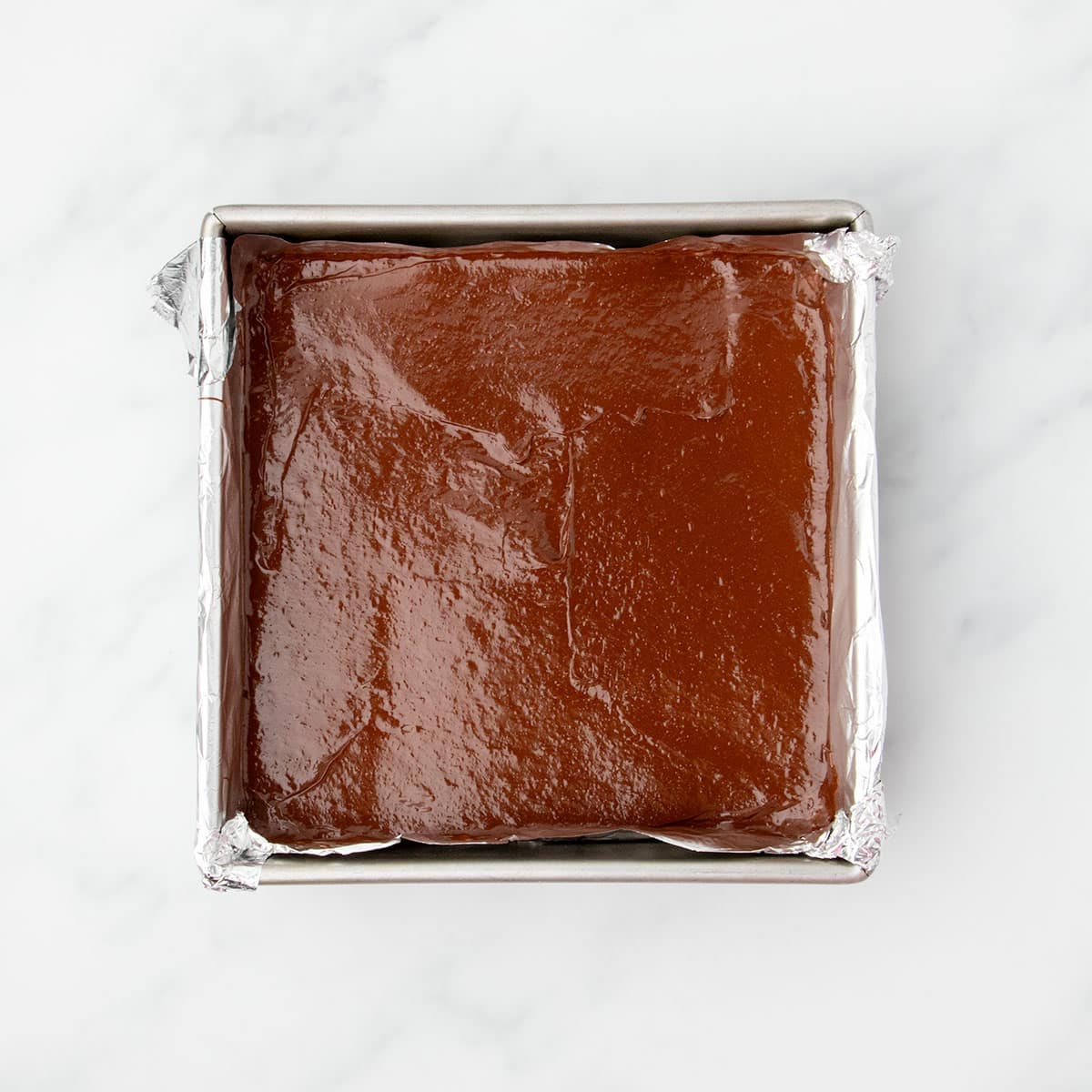 Unbaked brownie batter spread evenly in a foil-lined square metal pan