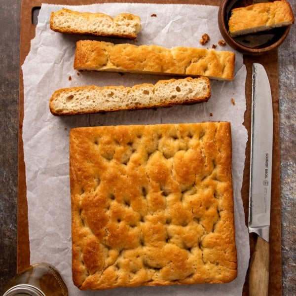 Rectangular loaf of golden flat bread on parchment paper, with three thick slices cut from the top and arranged above the loaf. A serrated bread knife rests alongside, and a small wooden bowl holds one slice.