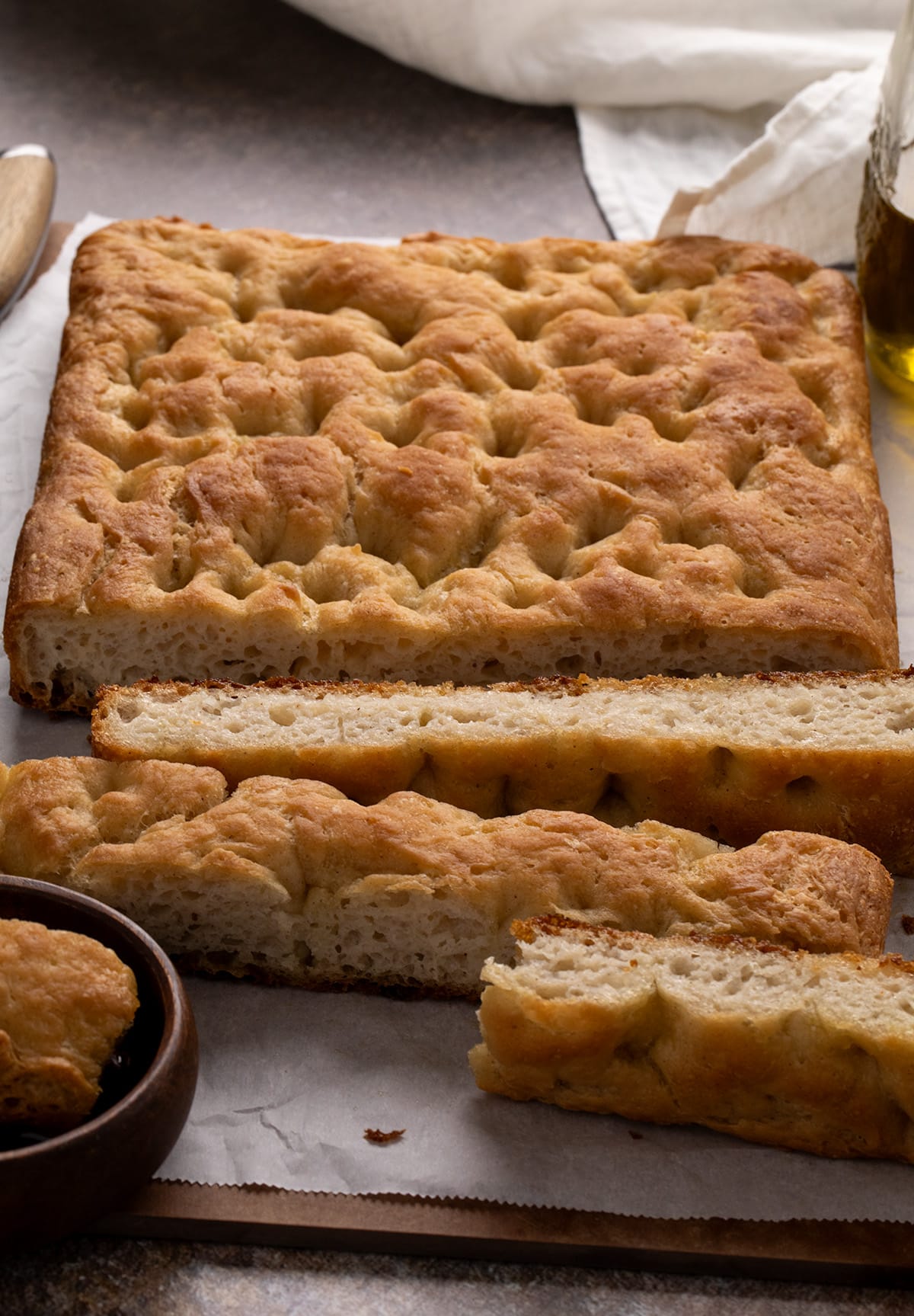 Side view of a rectangular bread loaf with a light, airy interior. Several thick slices rest in front of the loaf on parchment paper, with a white cloth and bottle of oil in the background.