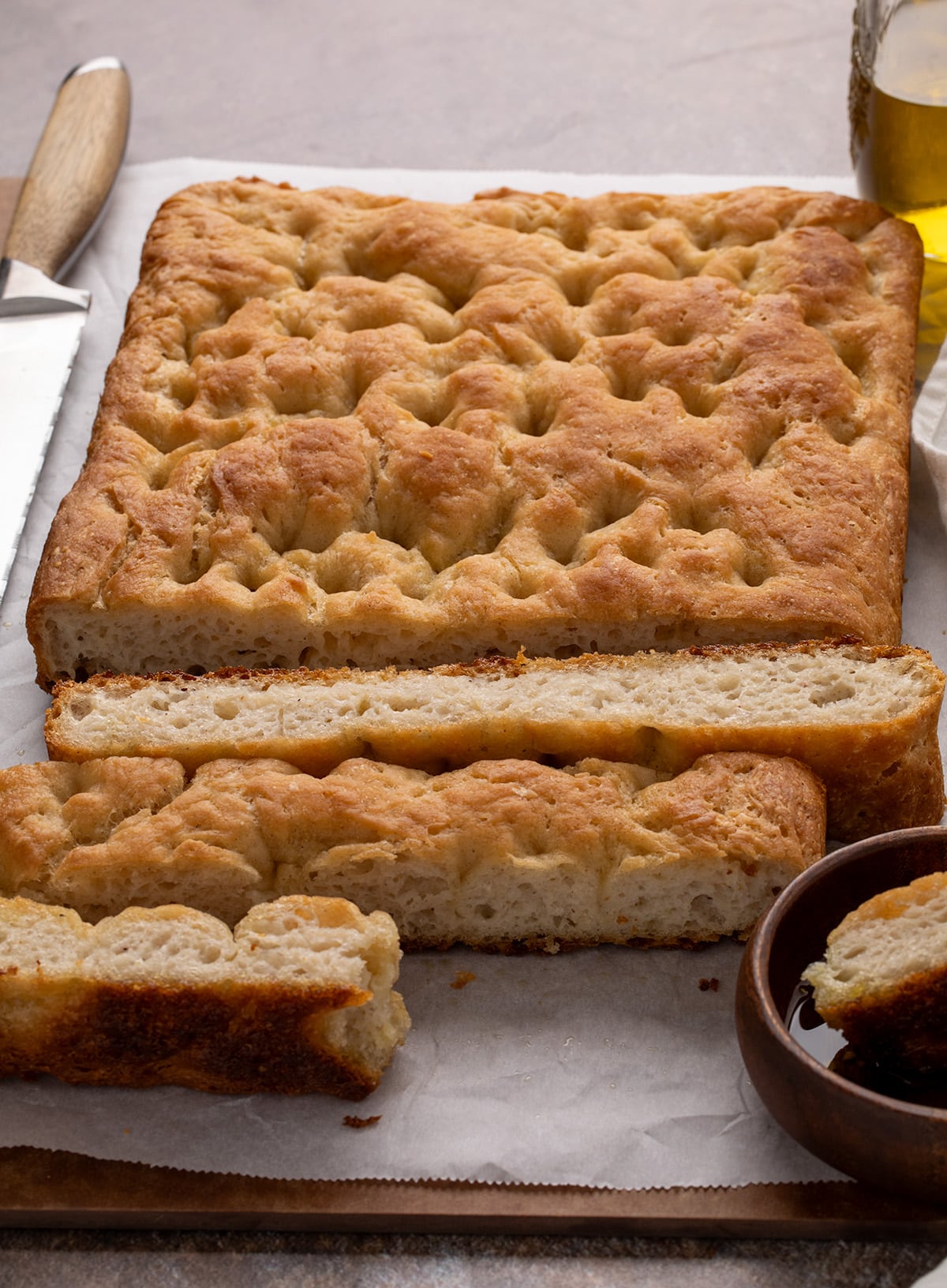 Angled view of a golden brown bread loaf with thick slices cut and resting in front, placed on a sheet of parchment with a serrated knife and a dish of oil partially visible.