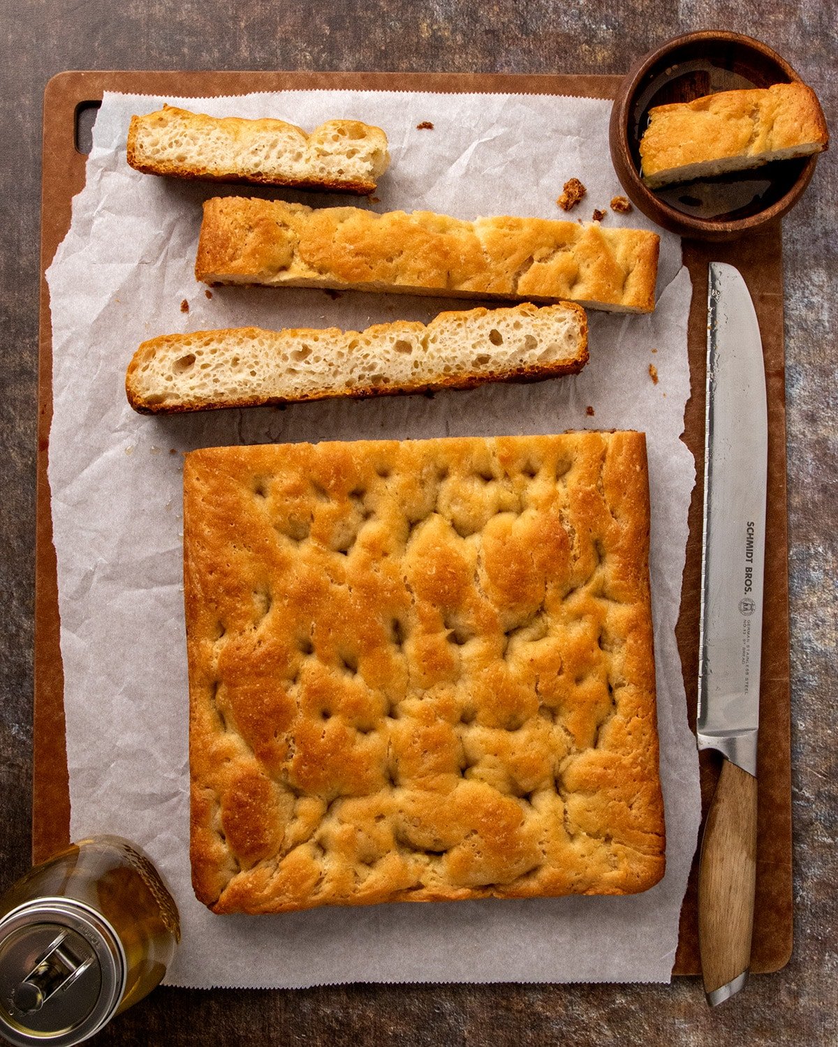 Top-down view of a golden gluten free focaccia loaf and three thick slices on parchment paper. A bread knife, glass bottle of oil, and wooden bowl with a bit of oil and a single slice frame the arrangement on a dark cutting board.