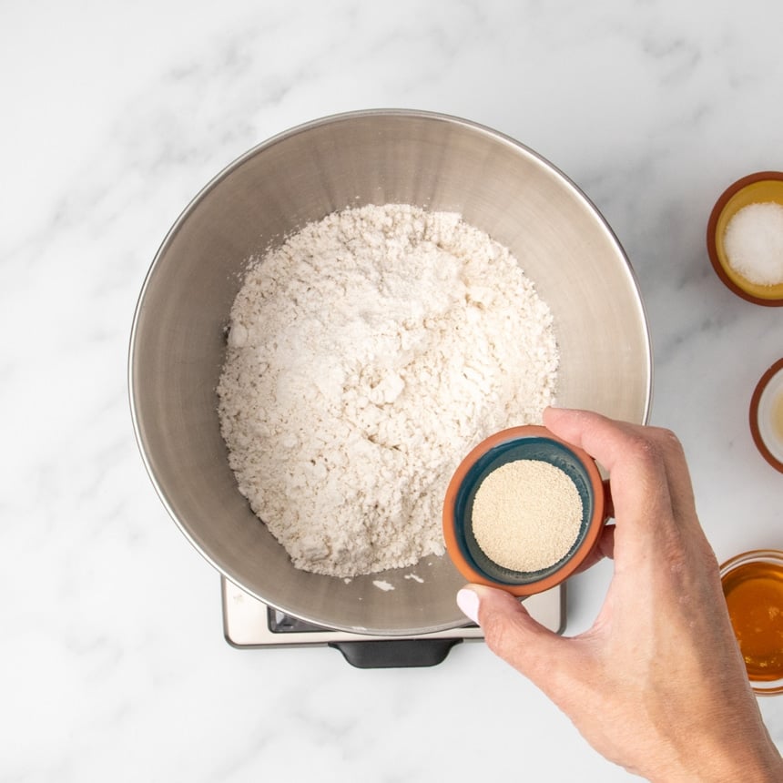 Metal mixing bowl with white flour mixture with hand tipping small pinch bowl of instant yeast granules into the bowl