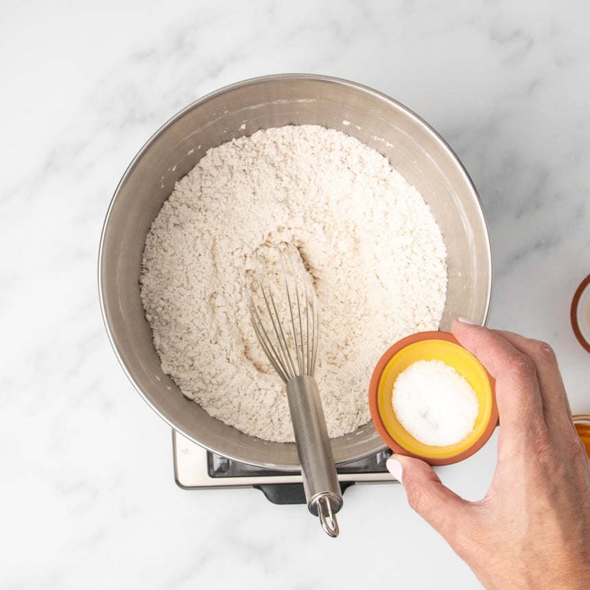 same mixing bowl, now with hand holding pinch bowl with coarse salt about to be added, with wire whisk in bowl