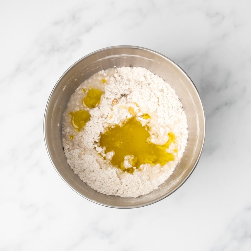 Metal round mixing bowl with dry flour mixture with olive oil, honey, water and vinegar added but not mixed in.