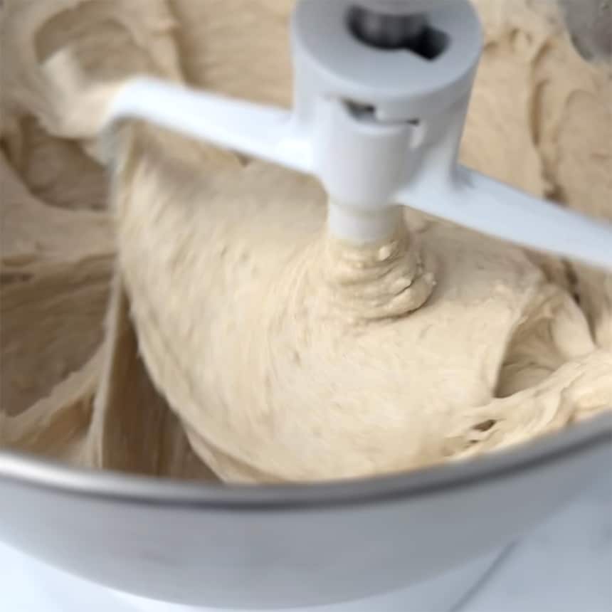 sticky, wet tan raw bread dough in metal mixing bowl being mixed with white paddle attachment
