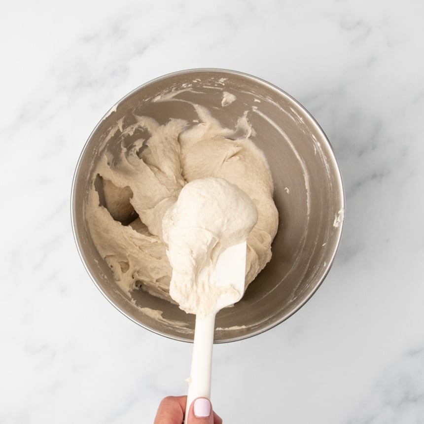Sticky, wet tan raw bread dough in mixing bowl with fingers holding white spatula with some dough on it