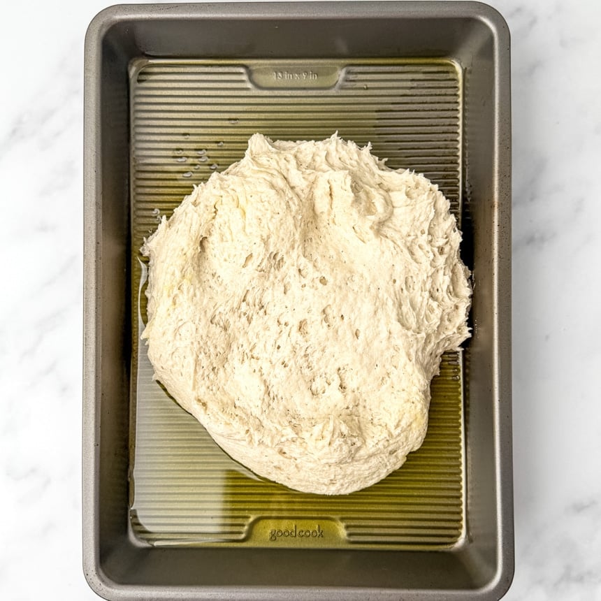 Pale raw dough in a mostly shapless mound resting in a rectangular metal pan with yellow olive oil on bottom