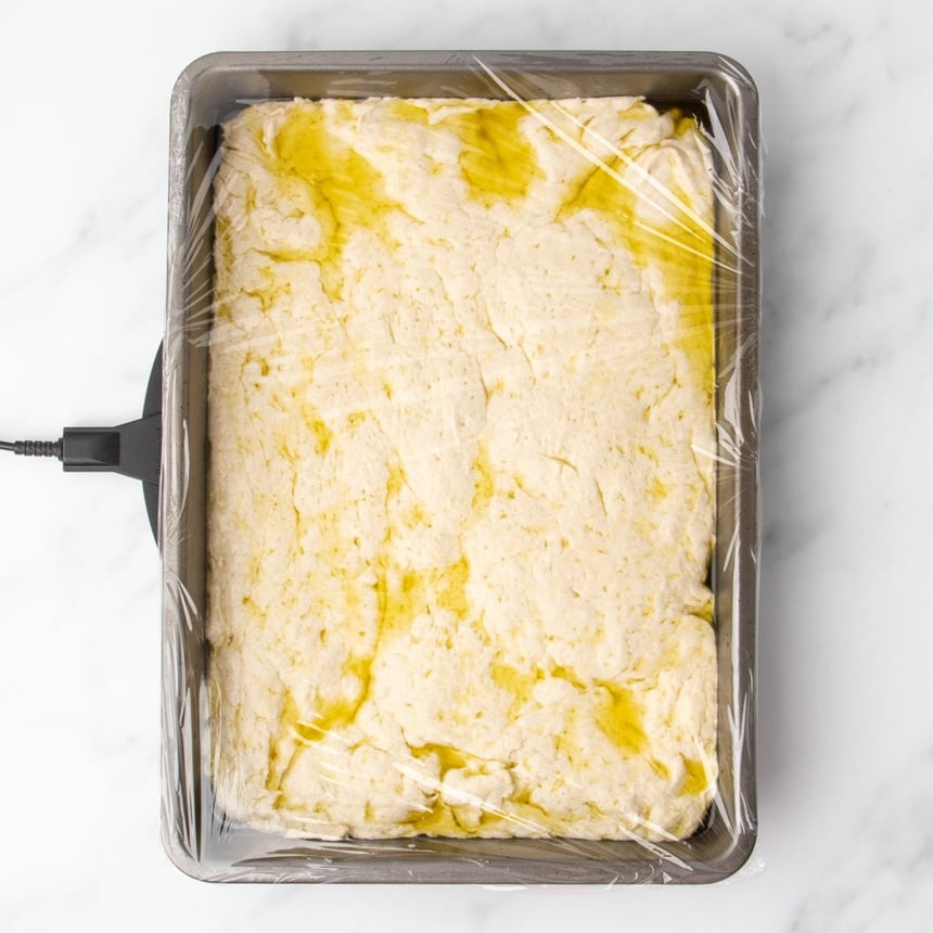 Pale dough shaped into a loose rectangle in baking pan with plastic wrap covering the top and pools of olive oil on top