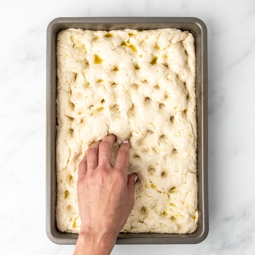 Fingers gently dimpling the surface of the risen dough in the baking pan