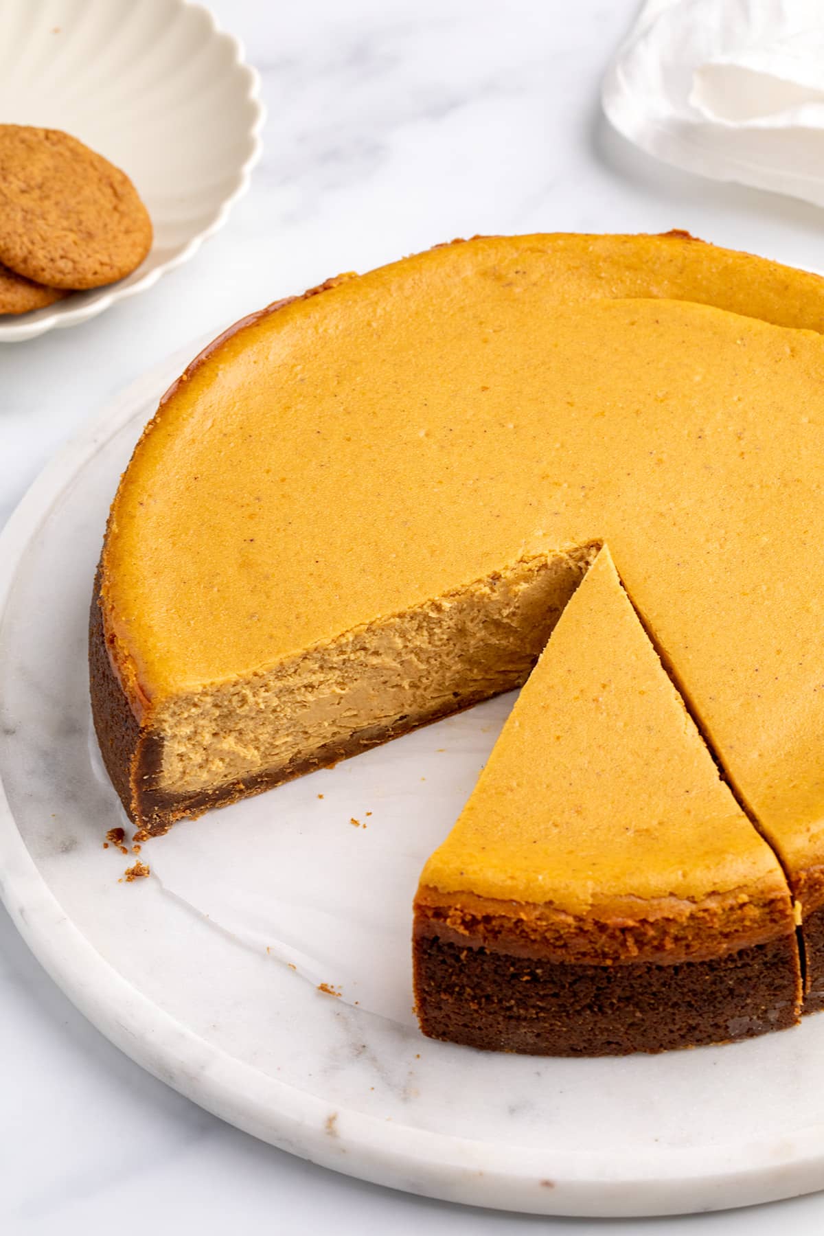 A whole baked cheesecake with a dark cookie crust on a marble platter, with one perfect slice cut and separated from the cake.