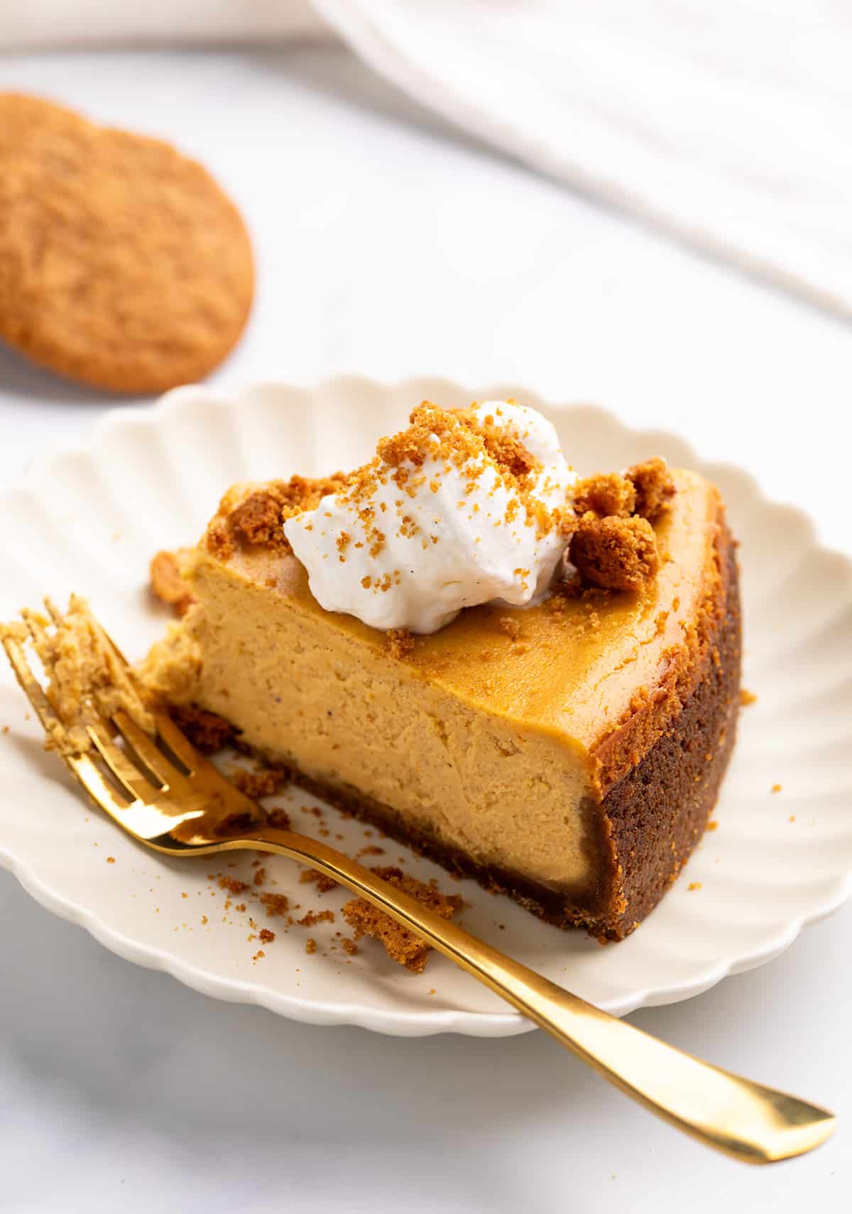 A side view of a slice of pumpkin spice cheesecake with a dark, thick crust, garnished with a dollop of whipped cream and cookie crumbles.