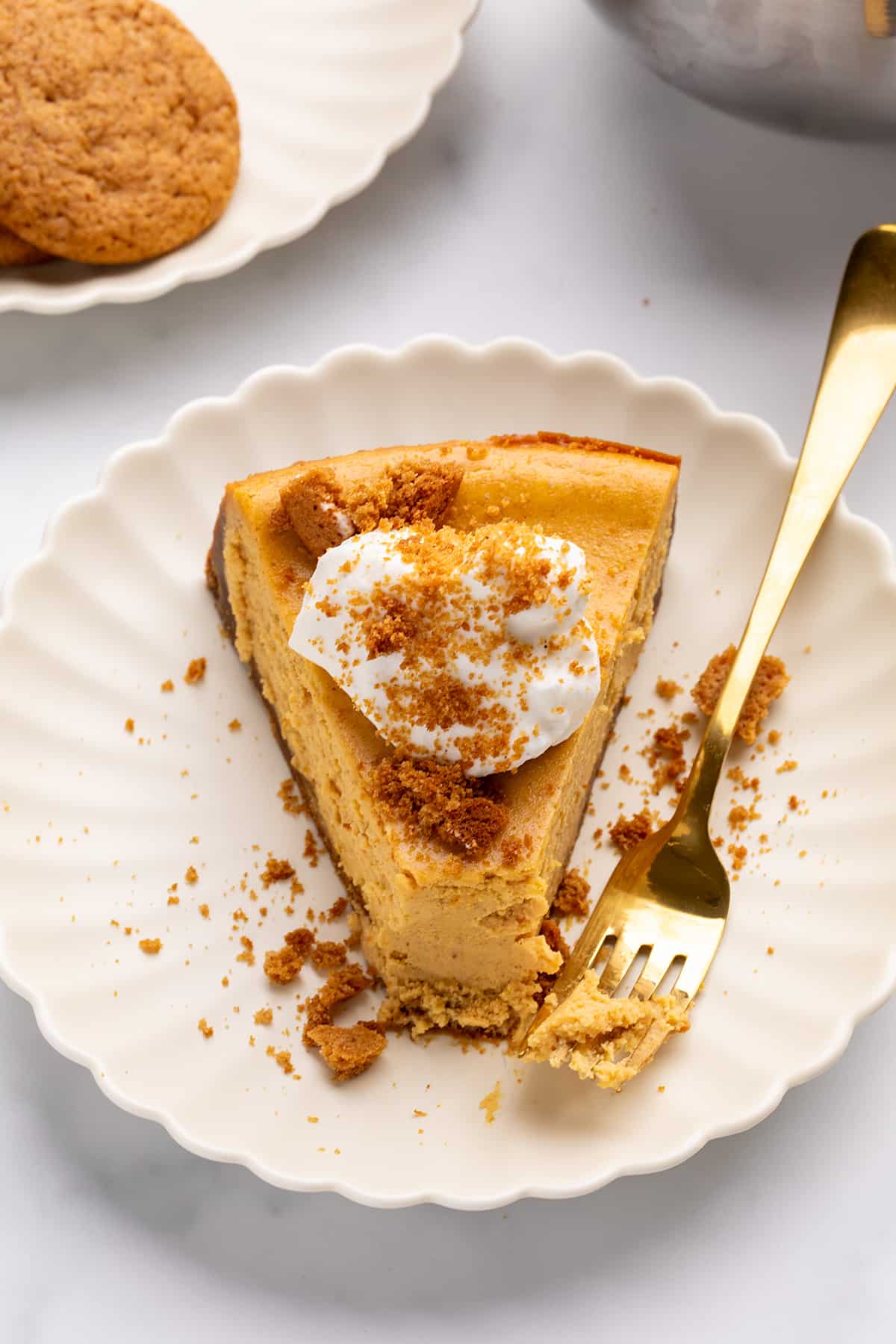 An overhead shot of a creamy slice of pumpkin cheesecake topped with whipped cream and crumbled cookie crust on a white plate, with a gold fork resting beside it.