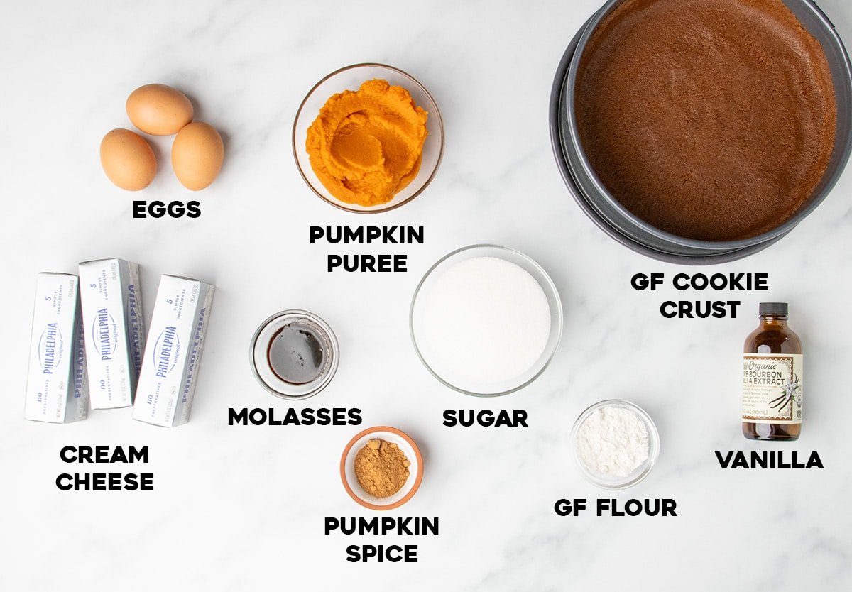 Labeled ingredients for a cheesecake recipe laid out on a marble surface, including cream cheese, pumpkin puree, sugar, eggs, and a prepared GF cookie crust in a springform pan.