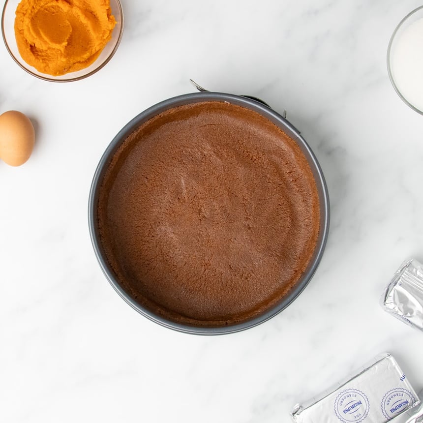 A springform pan with a dark, gluten free cookie crust pressed neatly inside, surrounded by other cheesecake ingredients on a marble counter.