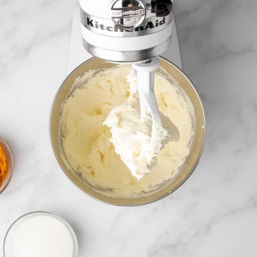 The paddle attachment of a stand mixer lifting up a scoop of fluffy, whipped cream cheese from the mixing bowl.