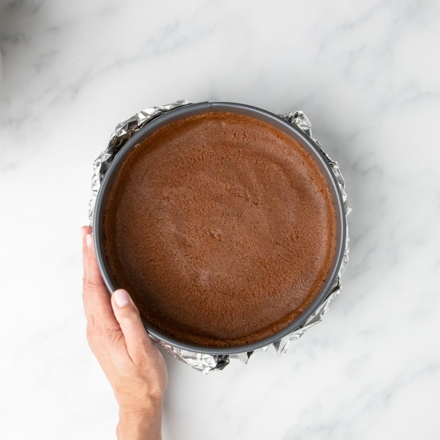 A hand holding a springform pan with a prepared cookie crust, now wrapped in aluminum foil to prepare it for baking.