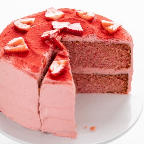 Two-layer pink frosted cake with two slices removed, topped with red powder and fresh strawberry slices.