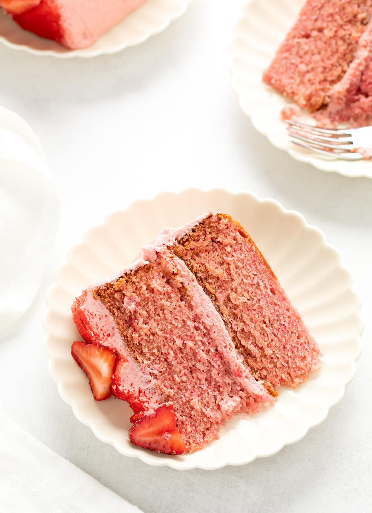 Slice of layered pink cake on a scalloped white plate, garnished with fresh strawberry pieces and two other plates with cake in background