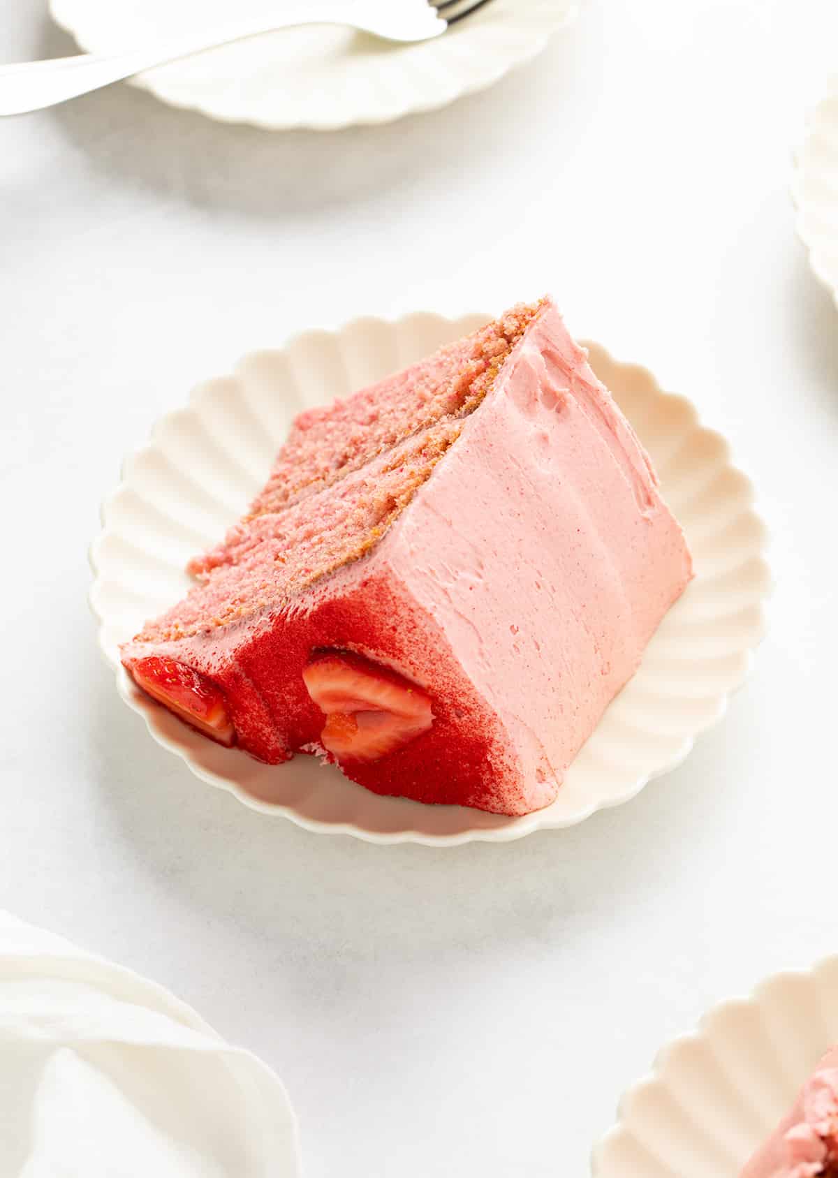 Close-up of a back and side of single cake slice on a white plate, showing thick frosting and strawberry pieces along the side.