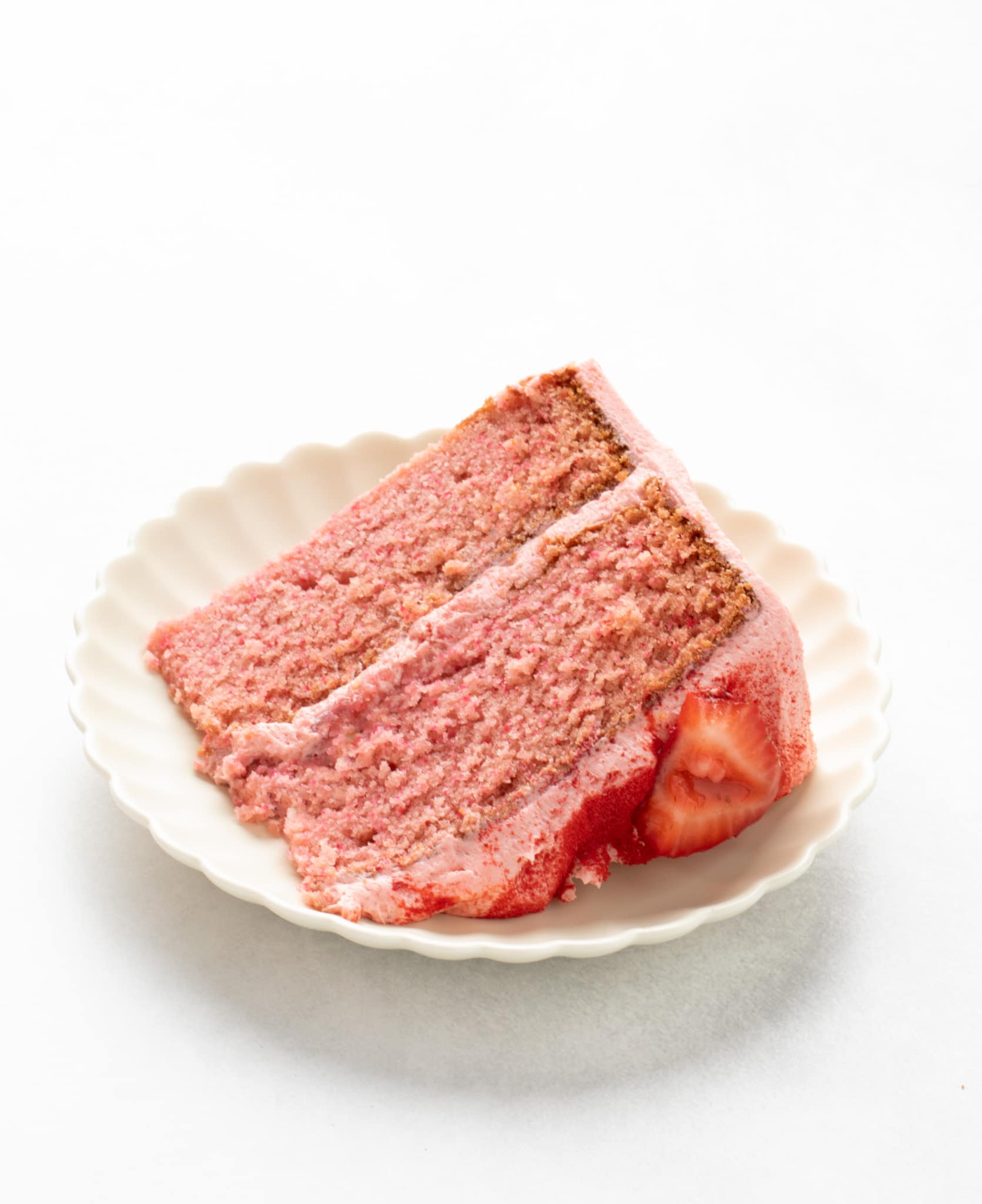 Two-layer slice of pink cake with strawberry pieces on the frosting edge, served on a scalloped plate.