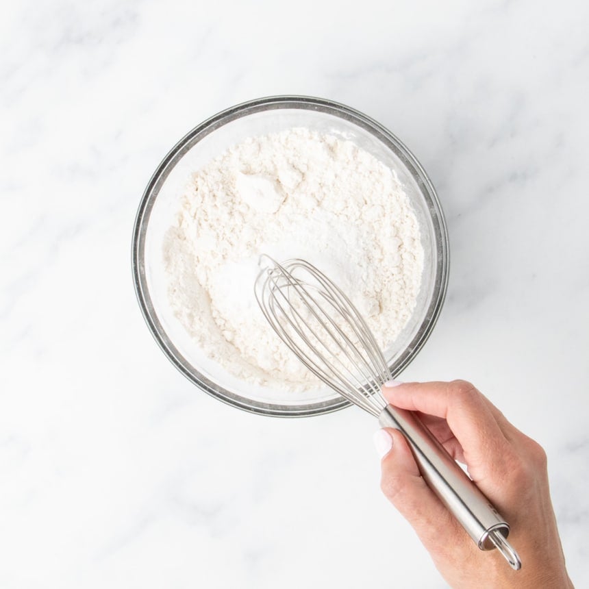 Hand whisking flour and leaveners together in a clear glass mixing bowl.