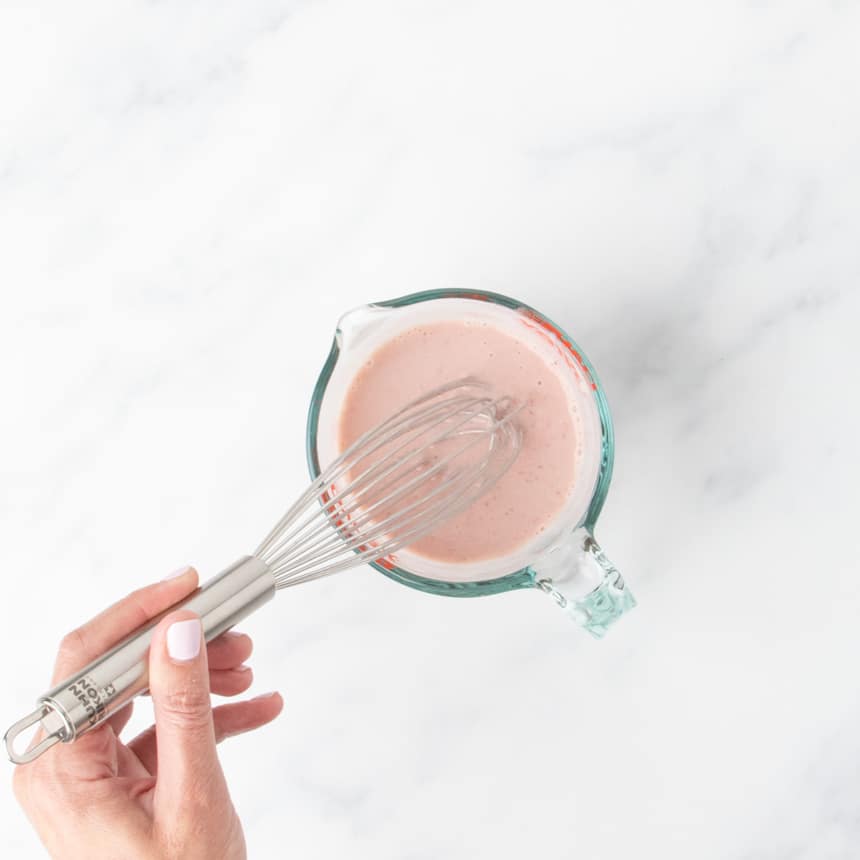 Pink liquid mixture in a glass measuring cup with a whisk inside